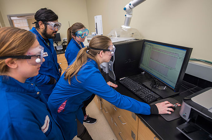 A group of chemistry students in protective goggles and suits study results on a computer monitor