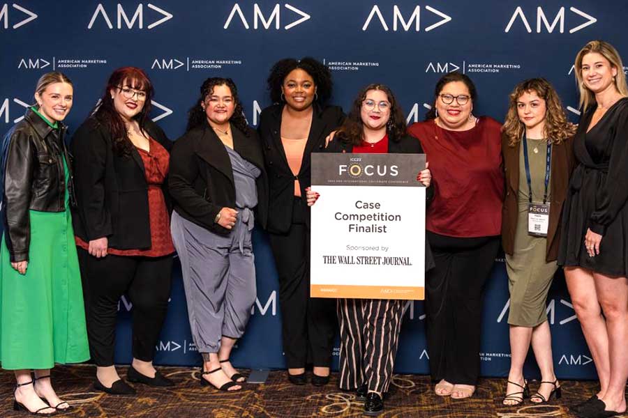 six AMA students stand with sign in front of AMA blue backdrop, accompanied by three staff members