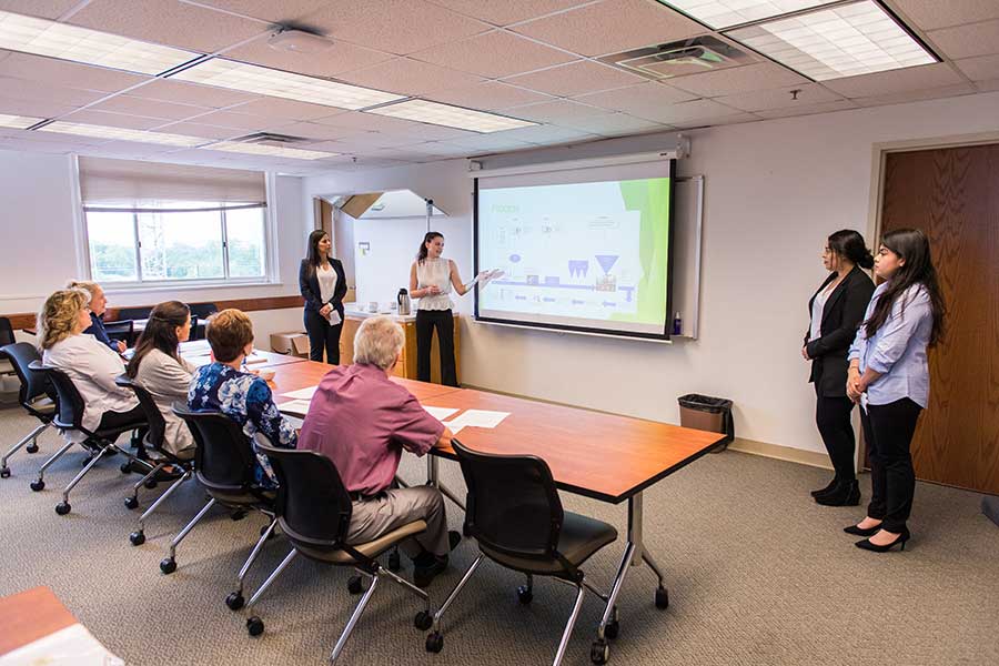 Three TWU female students in professional dress giving a presentation in a classroom setting.