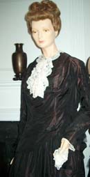 The long-sleeved black gown is shown on a mannaquin from the waist up. It has a white lace collar. 