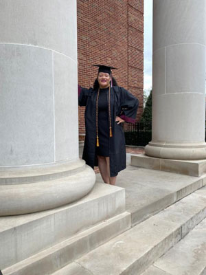Cheryl Brown leans against the a library column on TWU's Denton campus in her graduation gown.