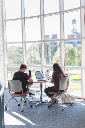 Two young women study at a table in Pioneer Hall with the Library visible out the window.