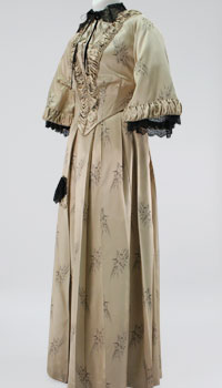 A soft beige brocaded dress with a long hoop skirt, fitted waist, and wide elbow-length sleeves. 