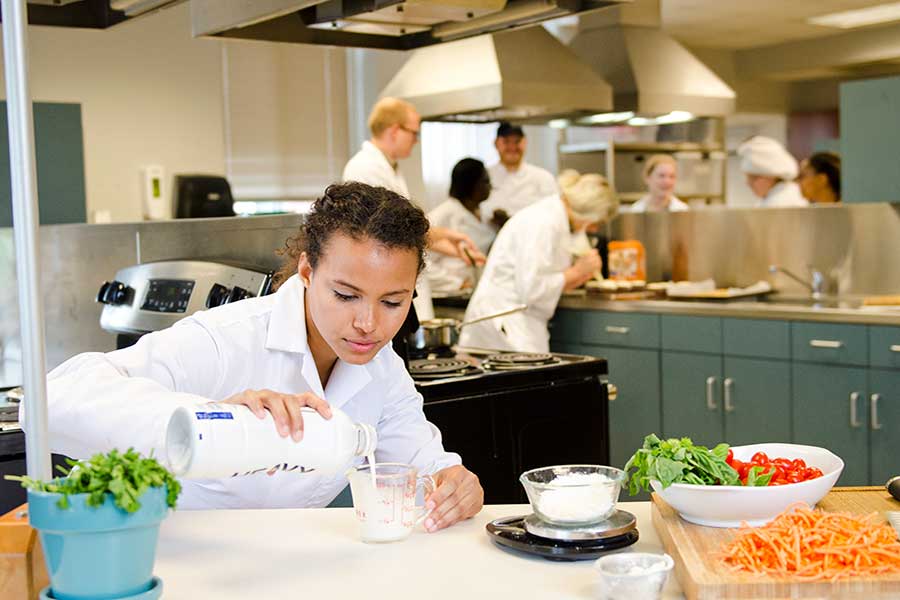 A TWU student measures milk into a cup while cooking in an industrial kitchen setting.	
