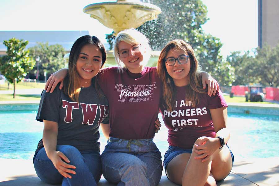 Three students in TWU shirts sit by the fountain on campus with their arms around each other.