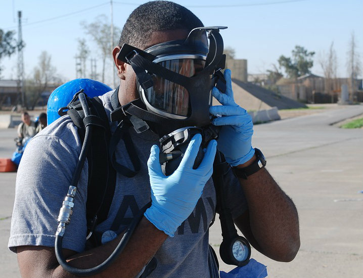A black male with a gray t-shirt and blue neoprene gloves donning a respirator.