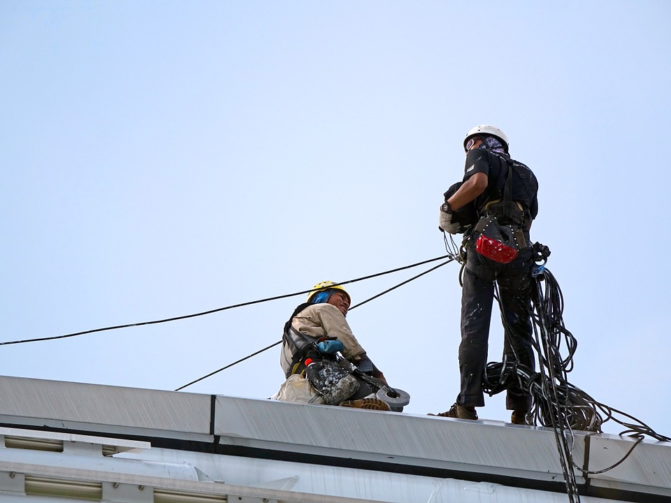Two employees using personal fall arrest systems and dressed in protective clothing, hard hats, and safety glasses on top of a building with a blue sky in the background.