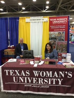 man and woman sitting and smiling at table draped with banner reading Texas Woman's University